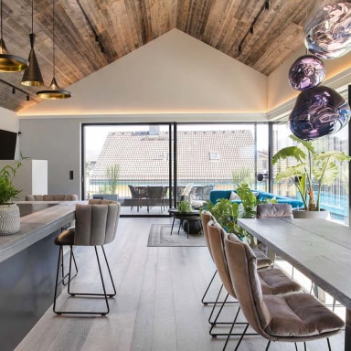 Offener Wohn- und Essbereich mit hoher Holzdecke, großen Glasfronten von Josko und Zugang zur Terrasse. Im Raum stehen ein Esstisch mit Polsterstühlen, moderne Hängelampen und viele Pflanzen, im Hintergrund ein blaues Sofa.