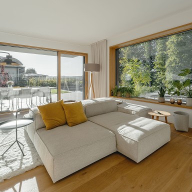 Heller, moderner Wohnraum mit bodentiefen Glasflächen und Ausblick auf Garten und Terrasse. Im Zentrum steht ein großes, helles Ecksofa mit zwei senfgelben Kissen, daneben kleine Beistelltische und ein flauschiger Teppich. Links führen große Schiebeelemente nach draußen, rechts sitzt ein breites Fenster von Josko mit Holzrahmen in der Wand, davor mehrere Grünpflanzen. Der Raum wirkt freundlich und natürlich durch viel Tageslicht und einen warmen Holzboden.