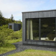 Detailansicht eines modernen Hauses mit dunkler Holzfassade und großem Holz/Alu-Fenster von Josko, umgeben von dichter grüner Vegetation.
