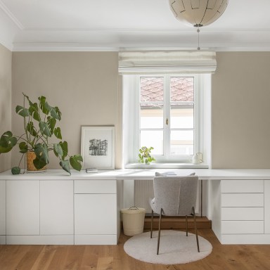 Modernes, helles Arbeitszimmer mit weißem Einbau-Schreibtisch, einem zentralen, klassischen Josko Fenster, einer Zimmerpflanze und einem schlichten Stuhl auf einem runden Teppich.