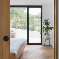 Modernes Schlafzimmer mit Holzparkett, großer Josko Terrassentür mit schwarzem Rahmen die zum Garten führt, einfachem Holzbett und Zimmerpflanze im Topf auf einem Ständer.