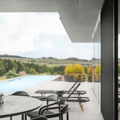 Überdachte Terrasse mit rundem Tisch und Stühlen im Vordergrund, zwei Sonnenliegen neben einem Infinity-Pool, große Glasfront auf der rechten Seite mit Spiegelung der Terrasse sowie weiter Blick über grüne Hügel, Felder und einen Wald im Hintergrund.