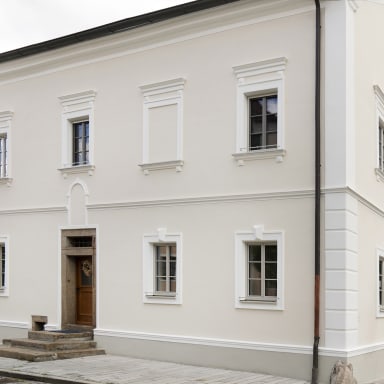 Frontansicht eines frisch sanierten Stadthauses mit heller Putzfassade, dekorativen Stuckrahmen und symmetrisch angeordneten, grau-weißen Fenstern von Josko im klassischen Stil.