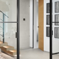 Detail eines modernen Innenraums mit Loft-Schiebetür aus Glas, im schwarzen Metallrahmen, offener Holz-Innentür und freitragender Treppe vor Betonwand.