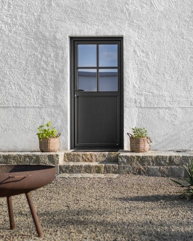 Schwarze Nebeneingangstür von Josko mit Sprossenverglasung in weiß verputzter Fassade, eingerahmt von Natursteinstufen und bepflanzten Körben.