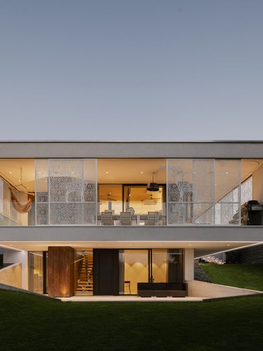 Modernes, zweigeschossiges Einfamilienhaus mit klarer, kubischer Architektur. Große, bodentiefe Glasflächen öffnen den Wohnbereich zur Umgebung. Im Obergeschoss befindet sich eine umlaufende Terrasse mit dekorativen, halbtransparenten Sichtschutzelementen. Warmes Innenlicht strahlt nach außen und schafft eine einladende Atmosphäre in der Dämmerung. Das Haus liegt leicht erhöht in einem grünen Hanggrundstück.