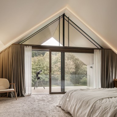 Helles Schlafzimmer mit großer Glasfront von Josko und Dreiecksfenster unter dem Satteldach, Blick ins Grüne; im Raum stehen ein Bett, ein Sessel und ein Teleskop auf dem Balkon.