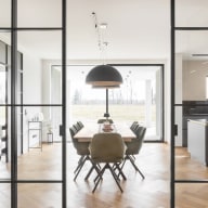 Blick durch eine raumhohe Loft-Schiebetür aus Glas und schwarzem Metallrahmen in einen hellen Essbereich mit Holztisch, Stühlen und großer Fensterfront.