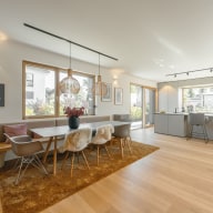 Großer, heller Küchen- und Essbereich mit Holzfußboden und großen Fenstern. Links steht ein langer Esstisch mit Stühlen und einer durchgehenden Sitzbank, dekoriert mit rosafarbenen Kissen und zwei hölzernen Pendelleuchten darüber. Rechts schließt die moderne, graue Küche mit breiter Arbeitsinsel, zwei Barhockern und einem großen Fensterband an. Die bodentiefen Schiebeelemente öffnen den Raum zum Garten, wodurch viel Tageslicht hereinfällt und eine warme, freundliche Atmosphäre entsteht.