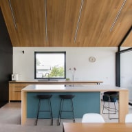 Moderne Küche mit Holzdecke, schwarzer Schrankwand, großer Kochinsel mit farbigem Akzent und weißer Arbeitsplatte, zwei schwarzen Barhockern und großen Glasflächen von Josko