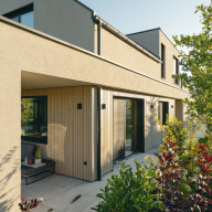 Seitenansicht eines modernen Einfamilienhauses mit sandfarbener Putzfassade und vertikalen Holzlamellen im Erdgeschoß. Große Schiebetüren und Fenster von Josko öffnen sich zum Garten, der mit dichtem Grün und rotlaubigen Sträuchern bepflanzt ist. Unter dem überdachten Bereich steht eine kleine Sitzbank mit Pflanzentöpfen. Der warme Abendlichtschein betont die klaren Linien und natürlichen Materialien der Architektur.