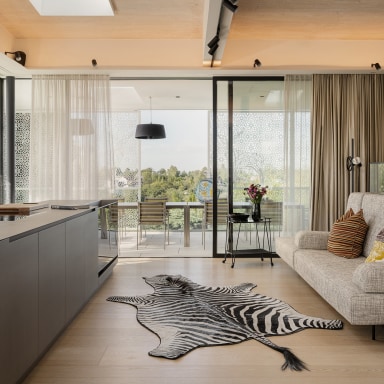 Offener, moderner Wohnraum mit integrierter Küche und großen Glas-Schiebetüren von Josko zur Terrasse. Eine Kücheninsel mit Spüle steht im Vordergrund, daneben ein Sofa mit Kissen auf hellem Holzboden. Vor den Glasflächen hängen leichte Vorhänge, dahinter ist ein Essbereich im Freien sichtbar. Ein dekorativer Teppich in Felloptik liegt zentral im Raum, warmes Tageslicht sorgt für eine helle, einladende Atmosphäre.
