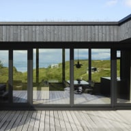 Große bodentiefe Fensterfront mit dunklen Rahmen öffnet den Wohn- und Essbereich zur Holzterrasse und zur grünen Landschaft mit Blick auf Wasser und Berge.