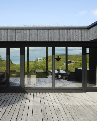 Große bodentiefe Fensterfront von Josko mit dunklen Rahmen öffnet den Wohn- und Essbereich zur Holzterrasse und zur grünen Landschaft mit Blick auf Wasser und Berge.