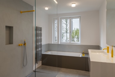 Modernes Badezimmer mit Dusche, Badewanne und Waschbecken; graue Flächen, gelbe Armaturen und Blick durch ein Josko Fenster ins Freie.