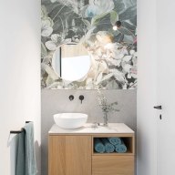 Modernes Gäste-WC mit floral gemusterter Wand, rundem Spiegel, weißem Aufsatzwaschbecken und Holzunterschrank.