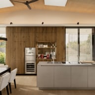 Großzügiger, moderner Wohn- und Essbereich mit offener Küche. Eine helle Kücheninsel mit integriertem Kochfeld steht im Mittelpunkt, dahinter eine warme Holzrückwand mit Einbaugeräten und offenen Regalen. Links befindet sich ein Esstisch mit gepolsterten Stühlen unter einer Hängeleuchte. Große Glasflächen und Schiebetüren von Josko lassen viel Tageslicht herein und verbinden den Raum mit dem Außenbereich.