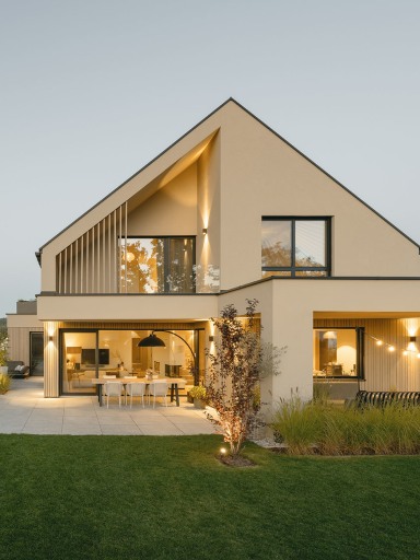 Modernes Einfamilienhaus in warmen Sandtönen mit großen Glasflächen und schlanken Fensterrahmen von Josko. Der Baukörper hat ein asymmetrisches Satteldach, raumhohe Fenster, vertikale Lamellen im Obergeschoß und einen offenen Essbereich, der durch große Schiebetüren zur Terrasse hin geöffnet ist. Im Vordergrund liegen ein gepflegter Garten und ein schmaler Pool, im Hintergrund Bäume und Nachbarhäuser. Abendliches Licht sorgt für eine warme, einladende Atmosphäre.