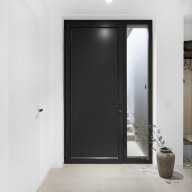 Minimalistischer Innenflur mit heller Wand und hellem Bodenbelag, mittig eine schwarze Nebeneingangstür von Josko mit schmalem, senkrechtem Glaseinsatz auf der rechten Seite, daneben eine bodenstehende Vase mit Zweigen, im Hintergrund ein offener Garderobenbereich.