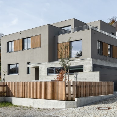 Modernes, zweigeschossiges Wohnhaus in kubischer Form mit grauer Fassade, großen Fenstern und Holzläden, davor ein Holzlattenzaun und gepflasterte Einfahrt mit Garage.