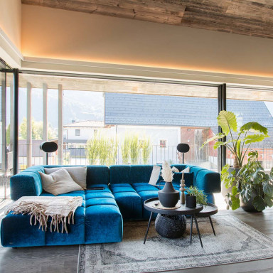 Wohnzimmer mit großem blauen Sofa, Couchtisch und Zimmerpflanzen vor bodentiefen Glasfronten von Josko. Durch die geöffneten Schiebetüren fällt Tageslicht in den Raum mit Blick auf Terrasse und Nachbarhäuser.