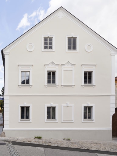 Frontansicht eines frisch sanierten Stadthauses mit heller Putzfassade, dekorativen Stuckrahmen und symmetrisch angeordneten, grau-weißen Holz-Alu- Fenstern von Josko im klassischen Stil.