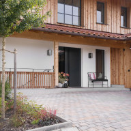 Eingangsbereich eines modernen Hauses mit Holzfassade, dunkler Haustür von Josko und großem Sprossenfenster darüber. Der Vorplatz ist gepflastert und mit kleinen Beeten und einem jungen Baum gestaltet.