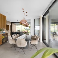 Großzügiger, heller Essbereich mit langem Esstisch und gepolsterten Stühlen, darüber mehrere kupferfarbene Pendelleuchten, links Holzsideboard vor Lamellenwand, im Hintergrund offene Küche, rechts bodentiefe Glas-Schiebetür von Josko mit Blick in den Außenbereich.