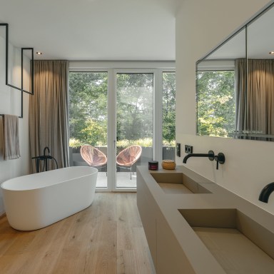Modernes, minimalistisches Badezimmer mit freistehender weißer Badewanne und bodentiefen Terrassentüren von Josko mit Blick ins Grüne. Rechts befindet sich ein breiter Waschtisch mit zwei rechteckigen Waschbecken und schwarzen Wandarmaturen, darüber ein großes Spiegelband. Links ist der Eingang zu einer Sauna zu sehen. Helle Vorhänge, warmes Holz am Boden und dezente Naturtöne verleihen dem Raum eine ruhige, elegante Atmosphäre. Auf der Terrasse stehen zwei runde Lounge-Stühle.