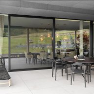 Überdachte Terrasse mit rundem Esstisch und mehreren schwarzen Stühlen aus Metall, bodentiefer Glasfront zum Wohnbereich im Hintergrund mit sichtbarem Esszimmer und warmen Pendelleuchten, links eine schwarze Sonnenliege, rechts Holzverkleidung an der Wand, Blick auf grüne Wiesen durch die Glasflächen.