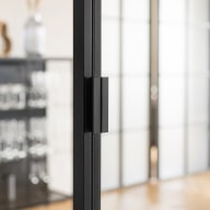 Detail eines schwarzen Griffs an einer Loft-Schiebetür aus Glas und Metallrahmen vor hellem Wohnbereich.