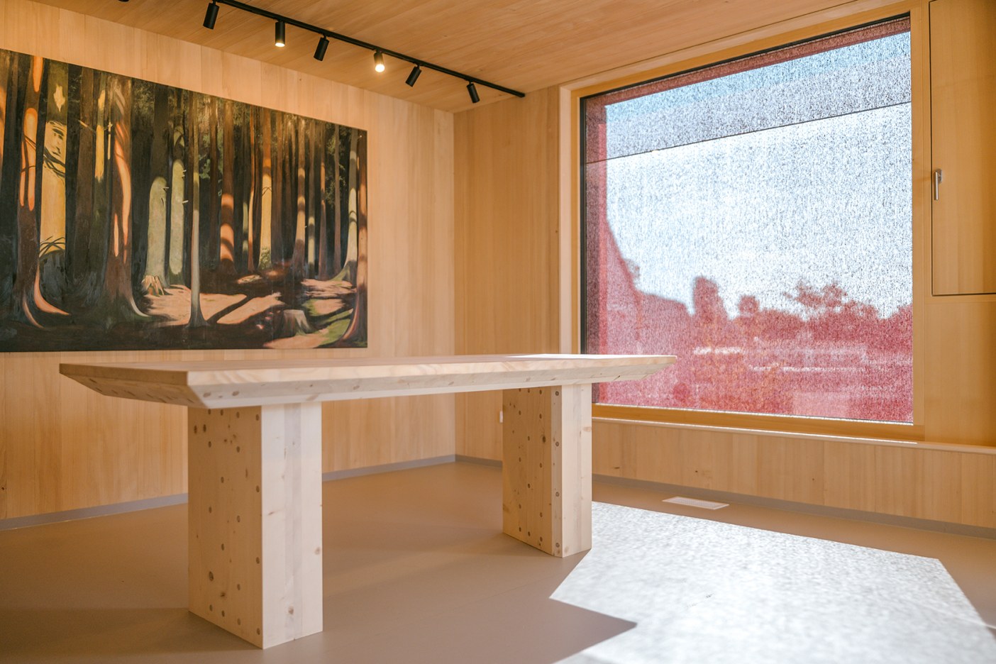 Innenraum mit hellen Holzoberflächen, einem massiven Holztisch und einem großen Fenster mit rotem Sonnenschutz von Josko. An der Wand hängt ein Gemälde eines Waldes, die Beleuchtung erfolgt über schwarze Deckenstrahler.