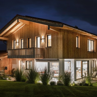 Stimmungsvolle Abendaufnahme eines modernen Holzhauses mit großen, leuchtenden Fensterflächen, die Wärme und Geborgenheit nach außen strahlen.