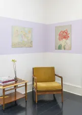 gallery installation of two drawings in the corner, with chair and end table with books a floral arrangement