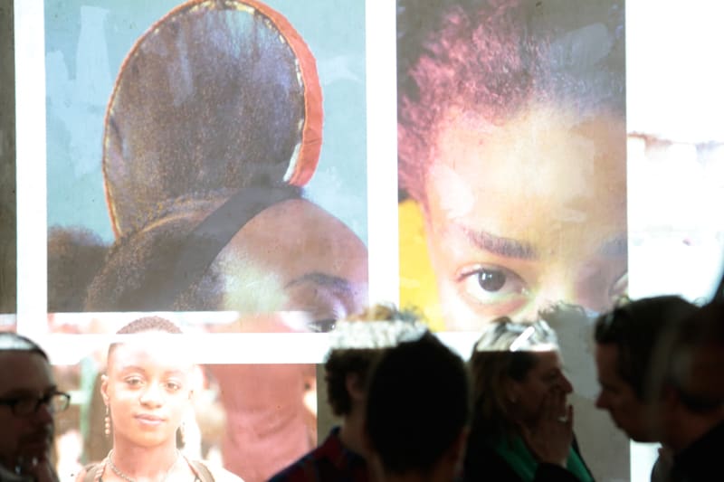 Wall projection of different women. Several exhibition visitors are talking in front of the wall.