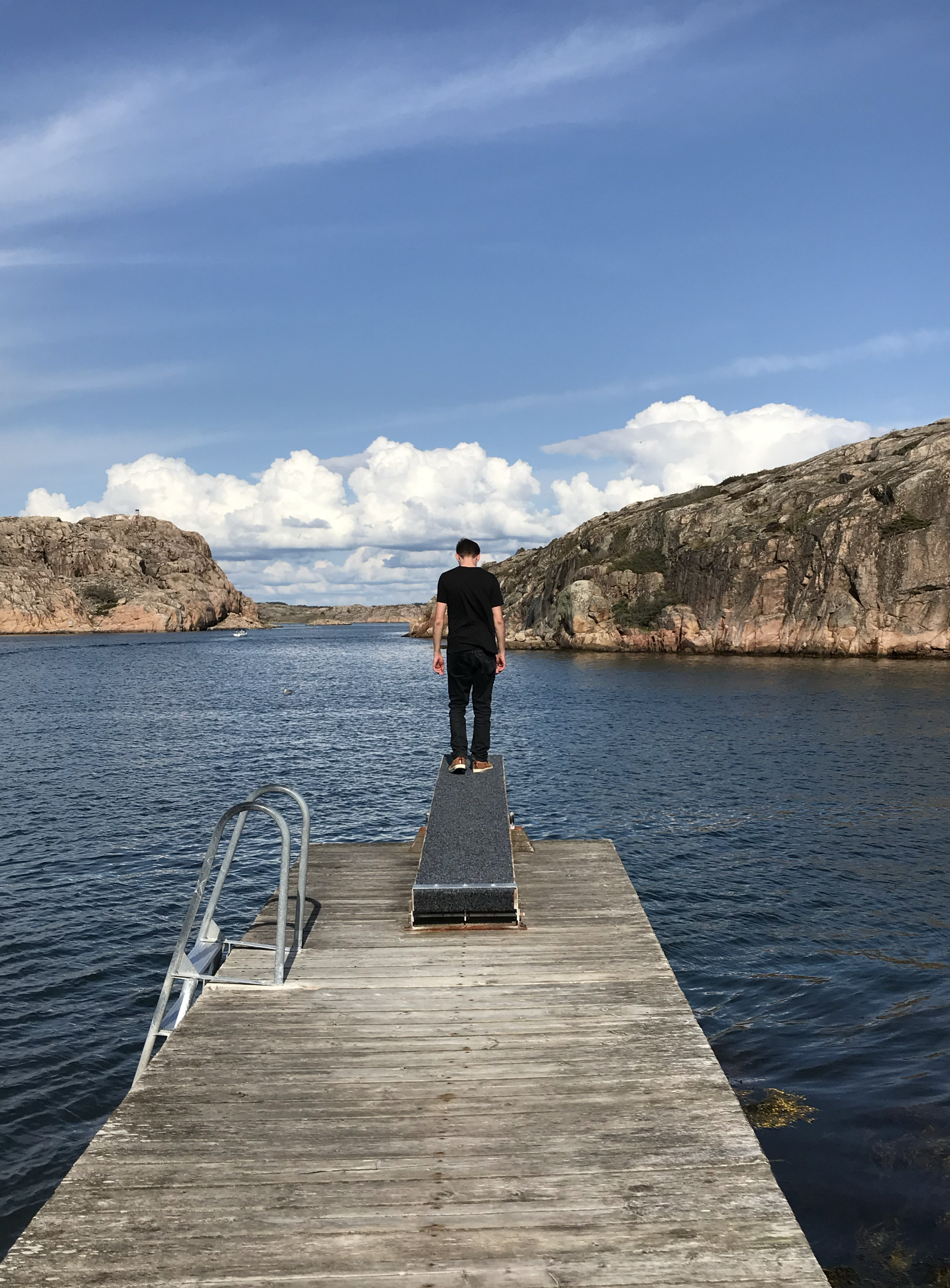 Steg mit Leiter ins Wasser und einer Sprungrampe, wo ein Mensch drauf steht und in das Wasser schaut. Im Hintergrund raken noch Felsen in das Bild.