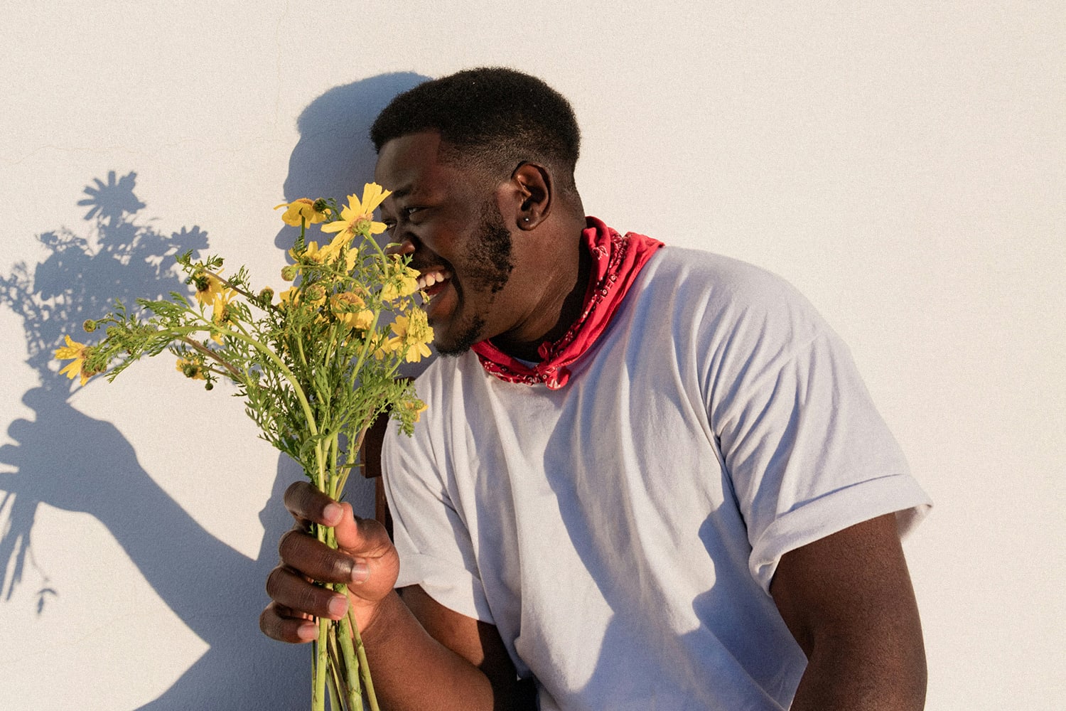 Ein Mann, der ein weißes T-Shirt und ein rotes Halstuch trägt, lacht, während er einen Strauß gelber Blumen vor einem schlichten weißen Hintergrund hält.