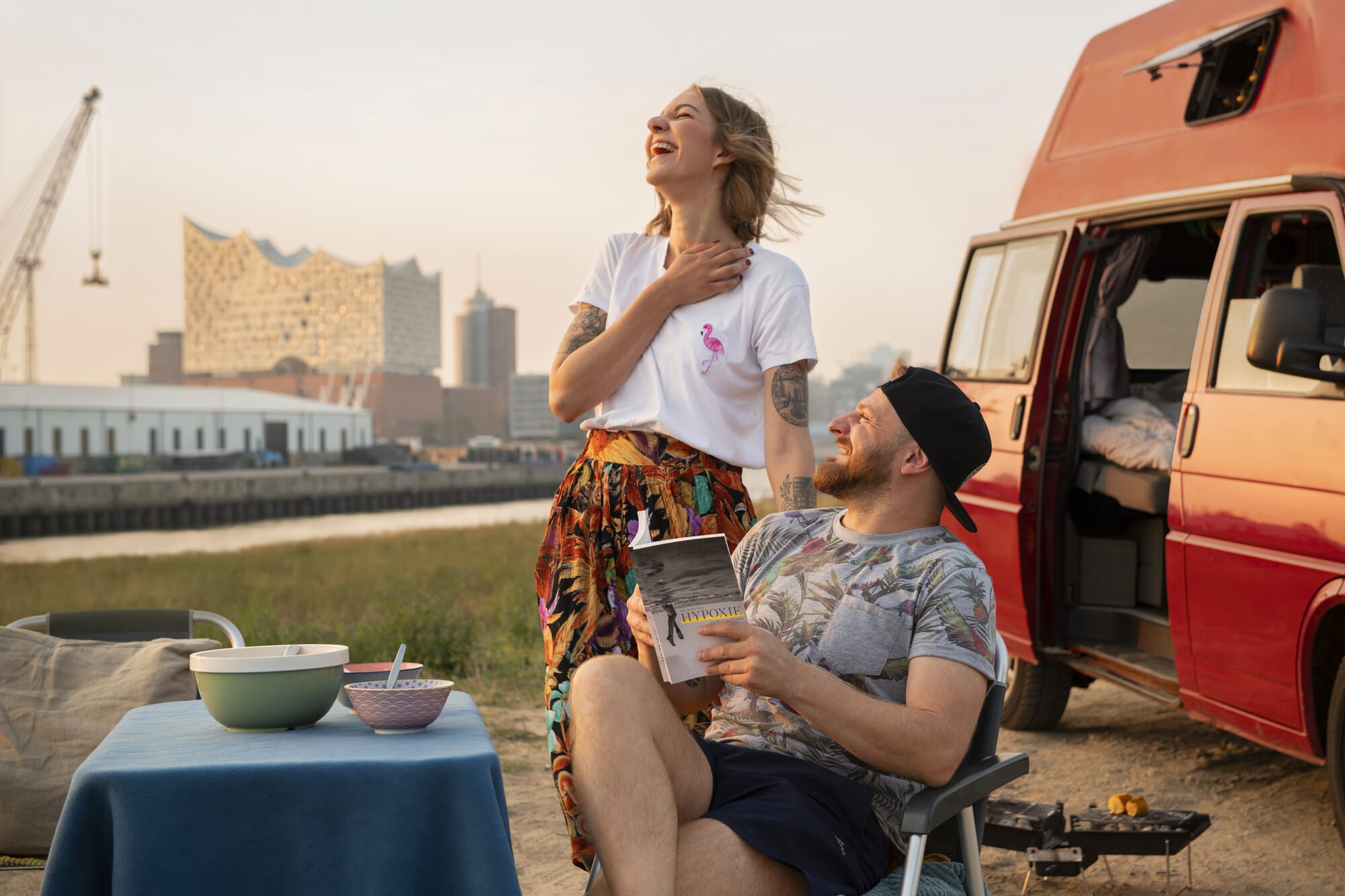 Kampagnenfotografie von zwei lachende Personen vor einem Karavan mit der Elbphilharmonie im Hintergrund. Eine Person sitzt und hat ein Buch in der Hand.