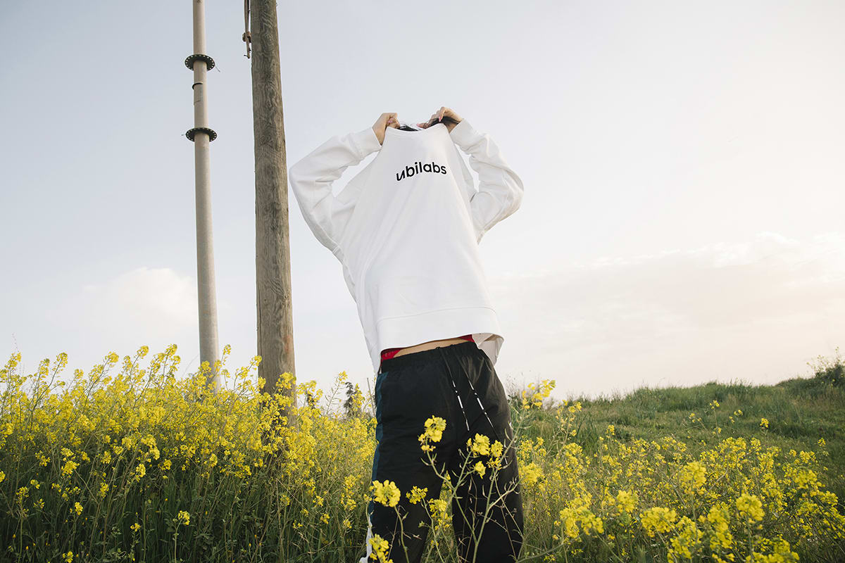 A person in a field of flowers, pulling a branded shirt over his head.