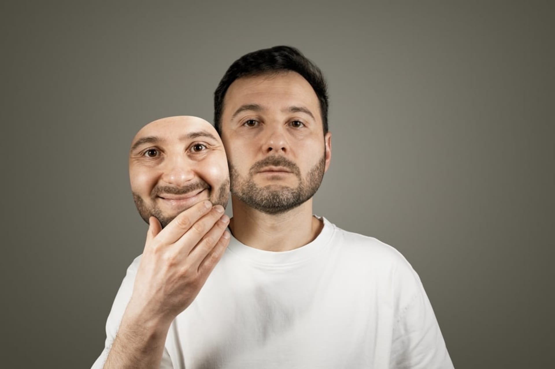A man in a white shirt holds a smiling mask in front of his face, revealing a neutral expression underneath.