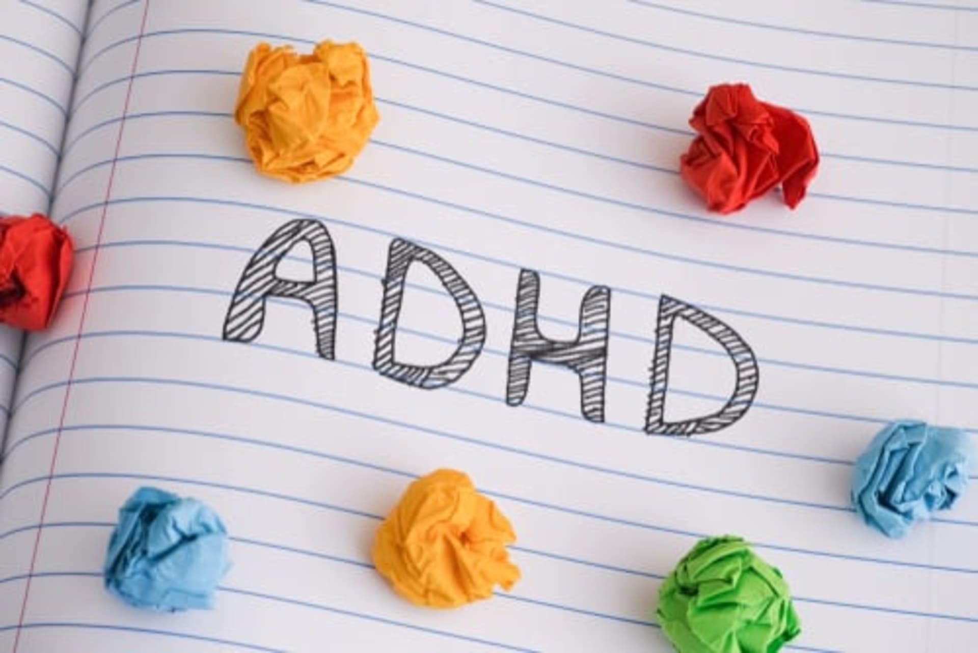 The word "ADHD" is written on a lined notebook page, surrounded by crumpled balls of colorful paper.