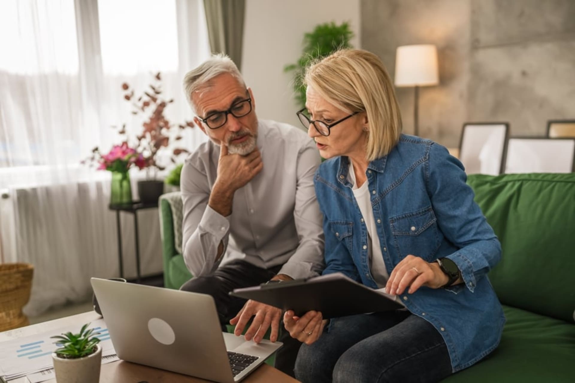 An older man and woman sit on a green couch, looking at documents and a laptop, appearing to discuss finances.