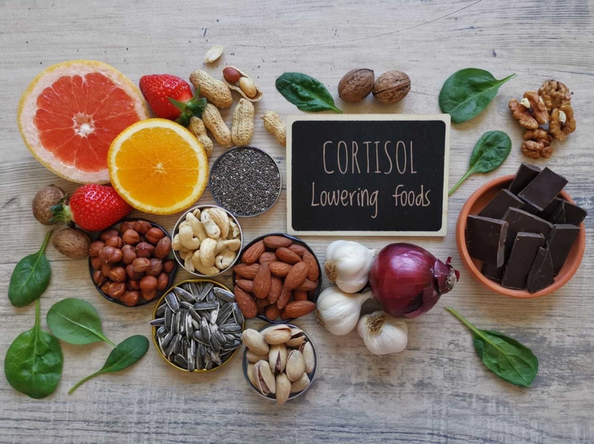 A variety of foods including citrus fruits, nuts, seeds, spinach, strawberry, garlic, onion, and dark chocolate surrounds a sign reading "CORTISOL Lowering foods."