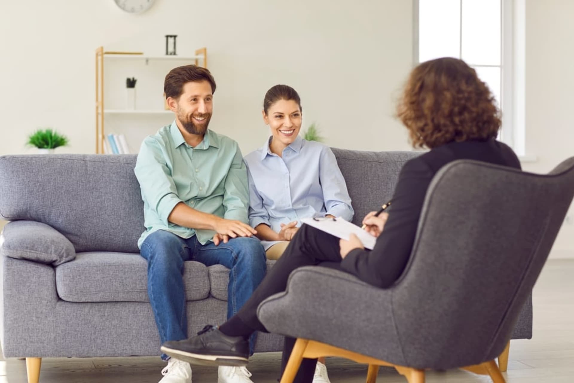 A couple sits together on a couch, smiling and holding hands, while talking to a professional during relationship counseling.