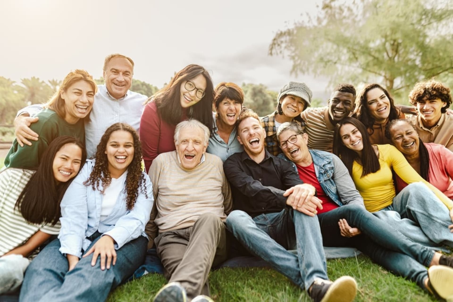 A diverse group of people of various ages sit closely together outdoors, smiling and laughing at the camera.