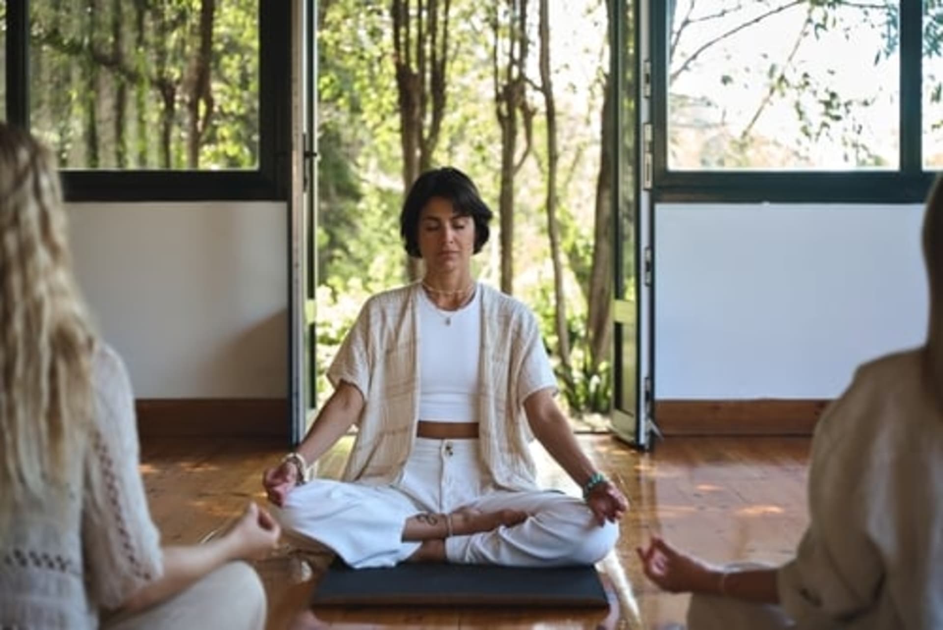 Three people sit cross-legged on mats in a calm, sunlit room with open windows, meditating together during a wellness retreat.