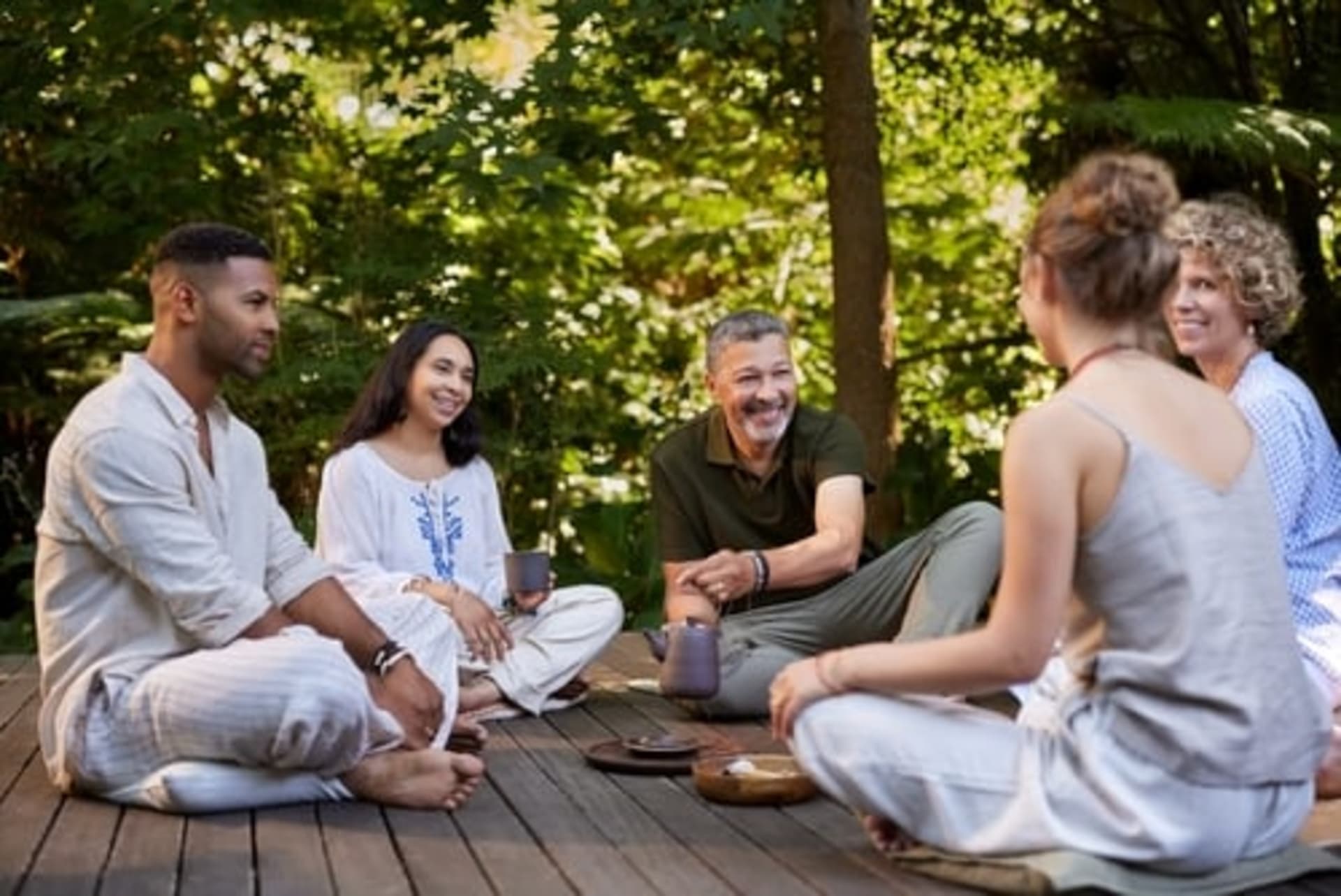 Five people sit cross-legged on a wooden deck outdoors, enjoying drinks and conversation amid lush greenery—a scene typical of wellness retreats.