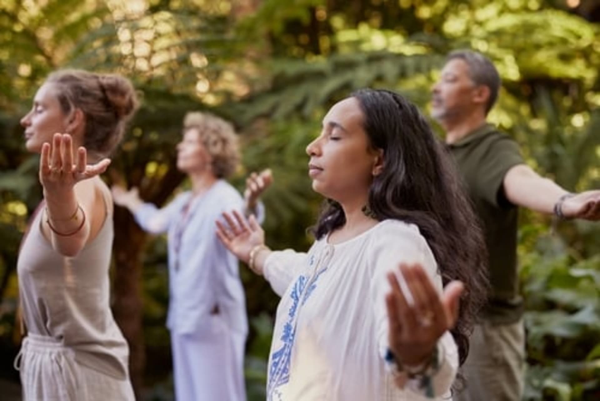 Four adults stand outdoors with eyes closed and arms outstretched, appearing to meditate or practice mindful breathing in a lush green setting.