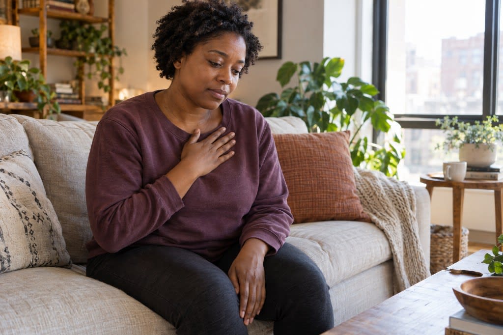 A woman sitting on her couch with one hand resting on her chest, offering herself warmth while a painful thought is present