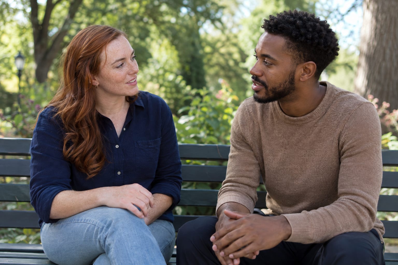 A red-haired woman and a Black man sit on a park bench facing each other and talking.